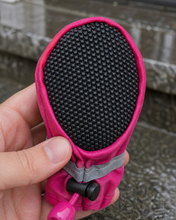A hand holds a pink dog boot showing its black textured grip sole.